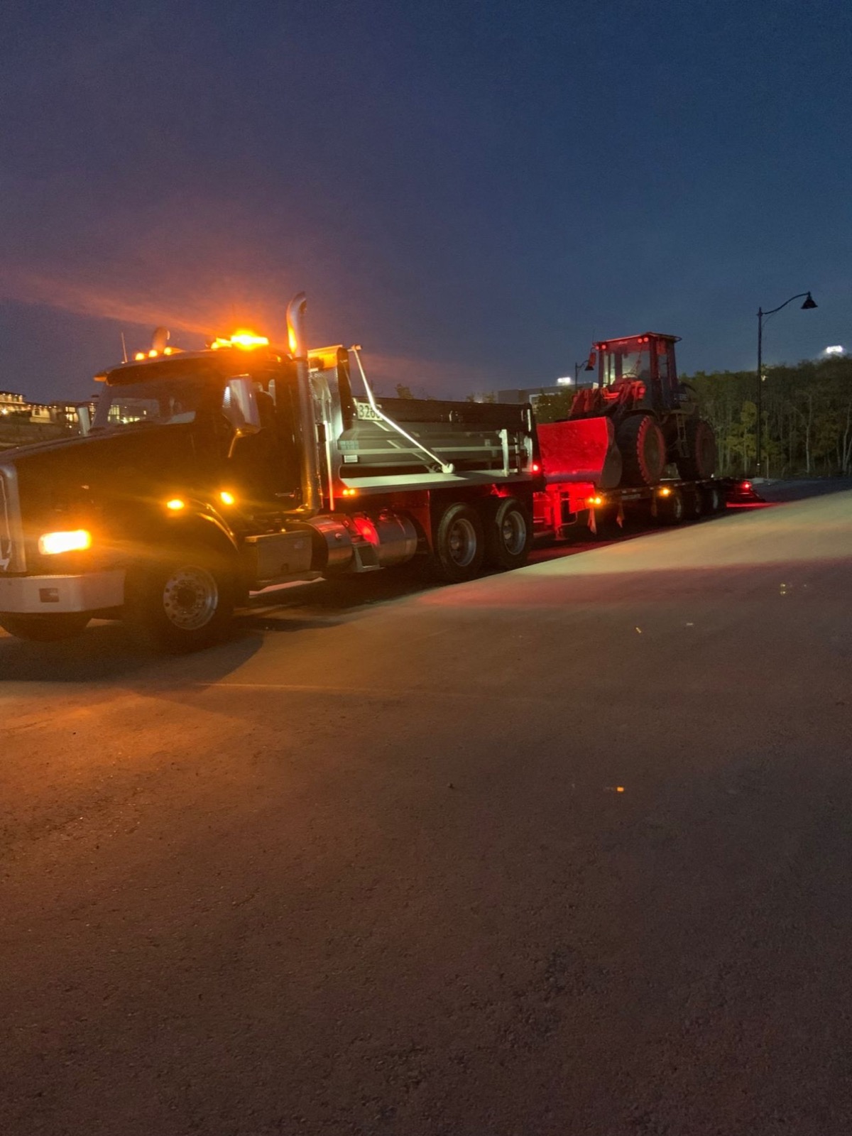 Dump truck with amber beacons hauling a loader at dusk